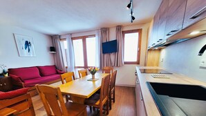 A compact living space with a dining area, a sofa, wooden chairs, a small kitchen with a sink, and a window with curtains.