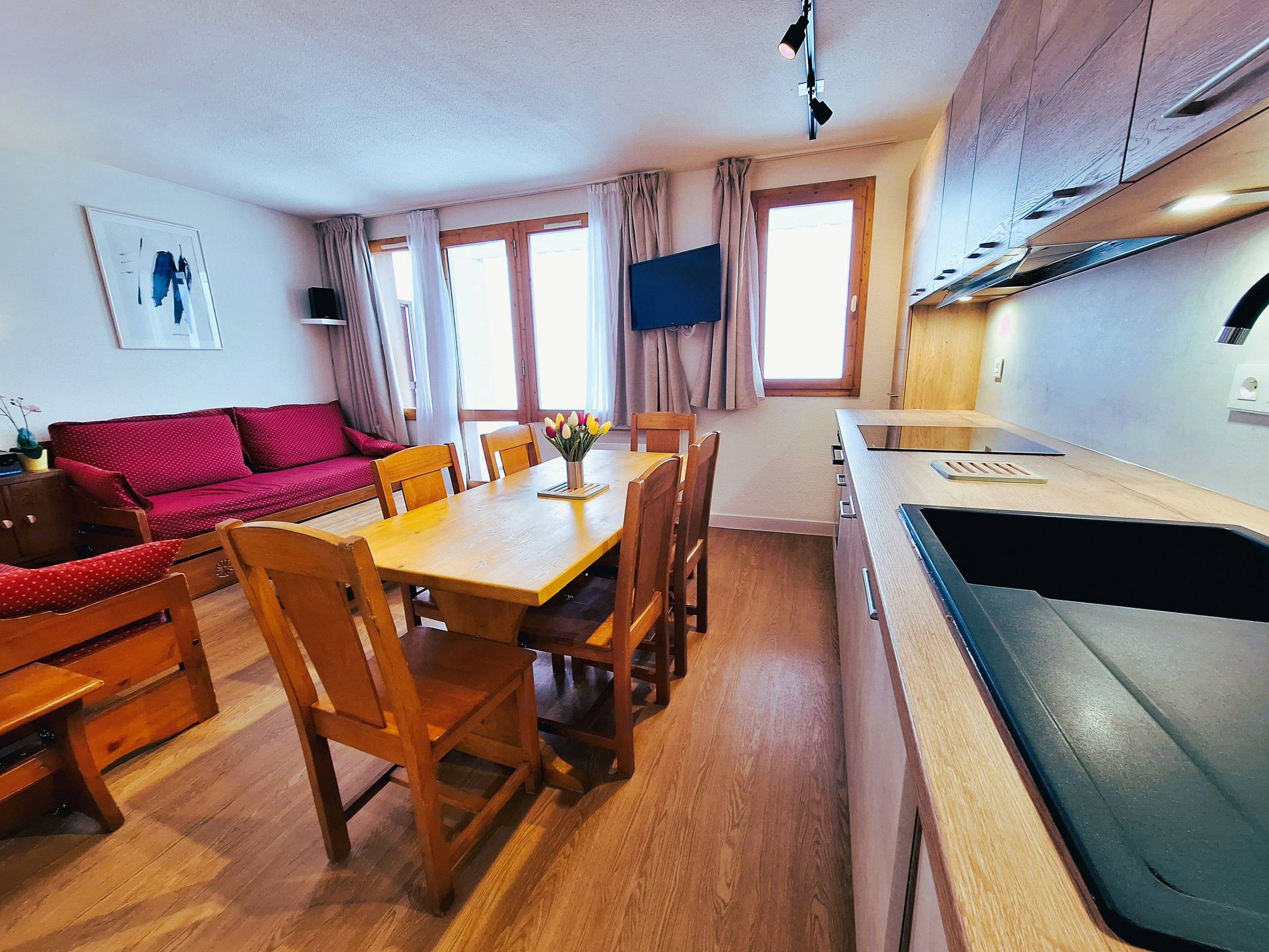 A compact living space with a dining area, a sofa, wooden chairs, a small kitchen with a sink, and a window with curtains.