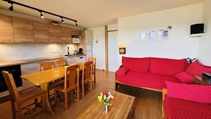 A compact living space with a wooden dining table, chairs, a red sofa, and a kitchen area with wooden cabinets and a microwave.