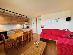 Um espaço compacto com uma mesa de jantar de madeira, cadeiras, um sofá vermelho e uma área de cozinha com armários de madeira e um micro-ondas.
