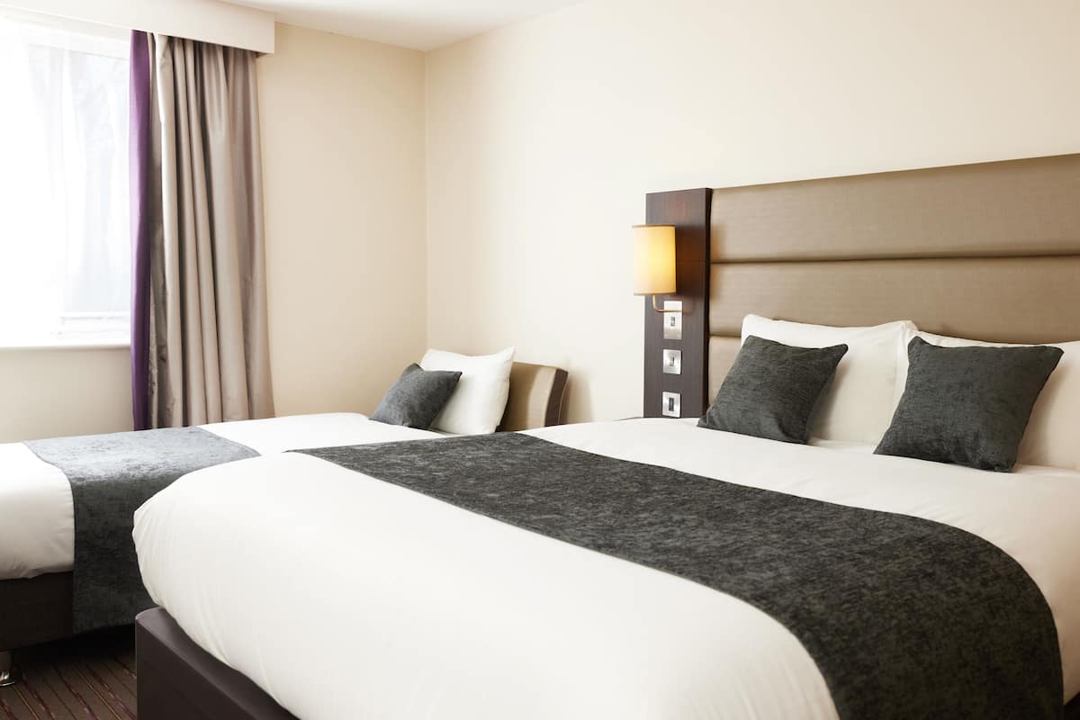 A hotel room with two beds, a headboard with a light fixture, and a window with curtains.