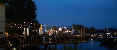 A waterfront area with a wooden bridge, string lights, and a calm water body reflecting the lights.