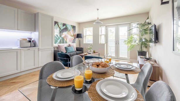 A modern kitchen with white cabinets, a dining area with a table set for breakfast, and a living room with a sofa and a large painting. - Modern 2 Bed Oasis -Parking & Garden - By Tempstay (Berkshire)