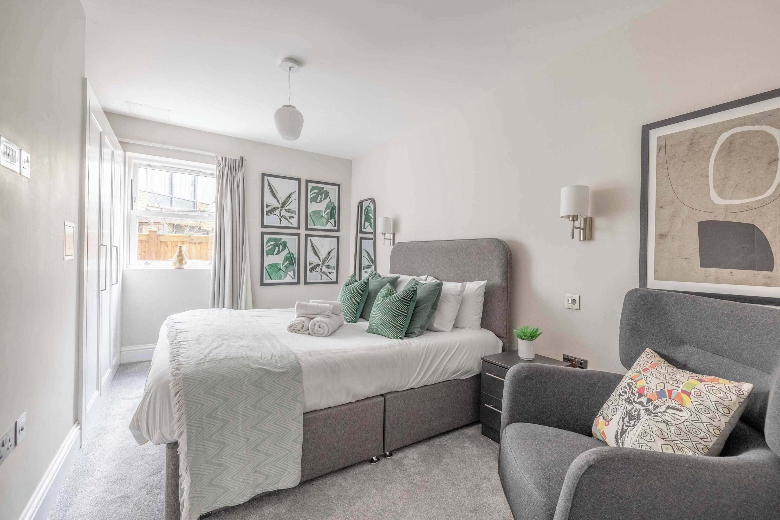 Een slaapkamer met een bed, een grijze fauteuil, een wandlamp en kunstwerken in lijsten.