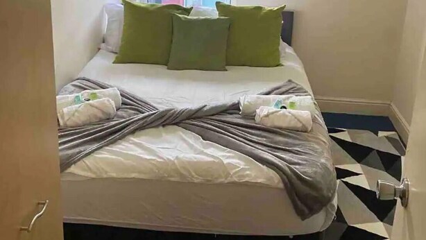 A small, neatly made bed with a white and gray duvet, green pillows, and a black headboard. A window with a view of a red building is above the bed. The floor has a black and white geometric pattern. - Flat 3, 52 Sneinton Hermitage Nottingham NG2 4BS (Nottingham)