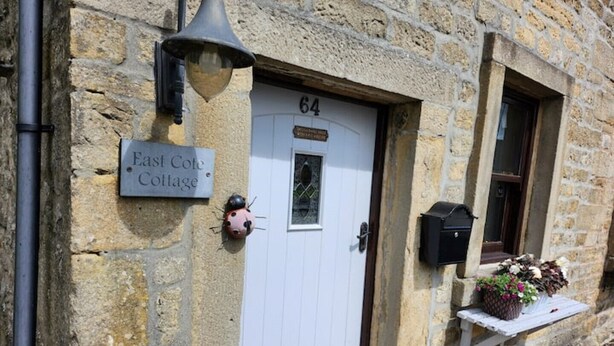 Une petite maison en pierre avec une porte blanche portant le numéro « 64 », une enseigne indiquant « East Cote Cottage » et une boîte aux lettres sur le côté droit.