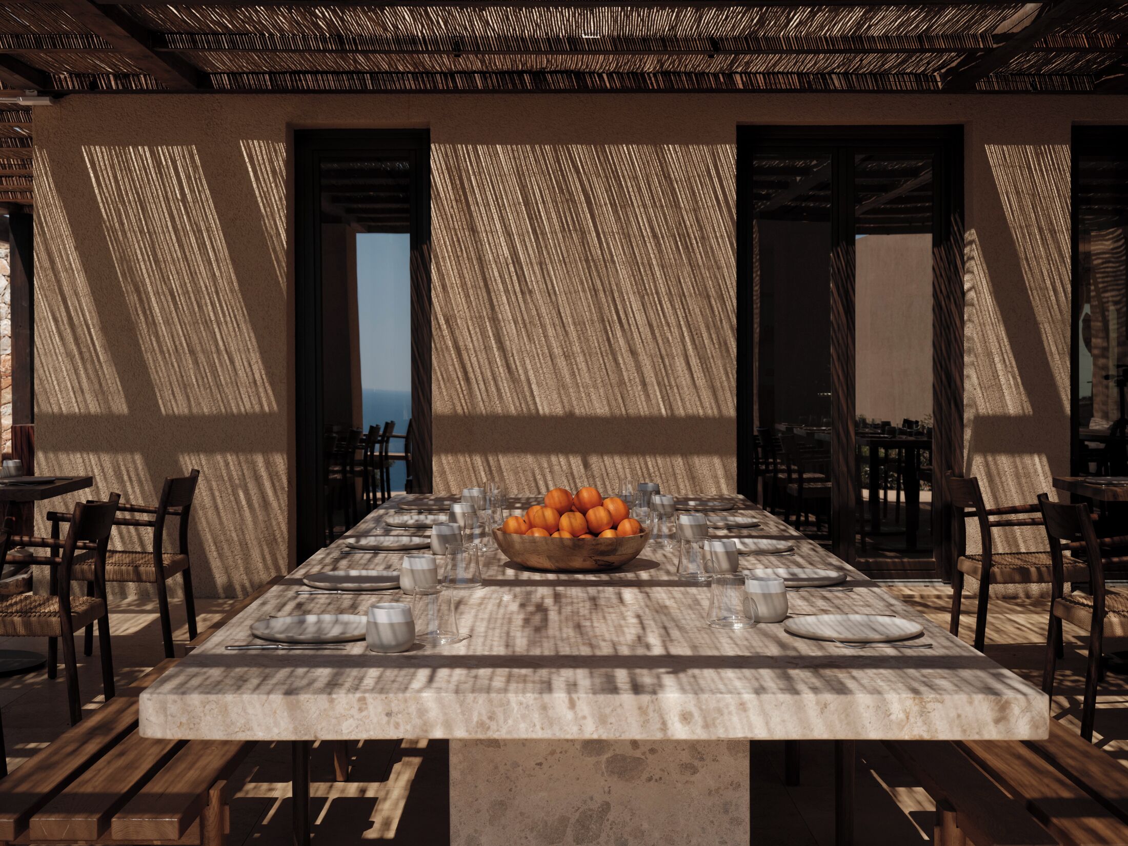 A long dining table set for a meal with a bowl of oranges, surrounded by chairs and plates.