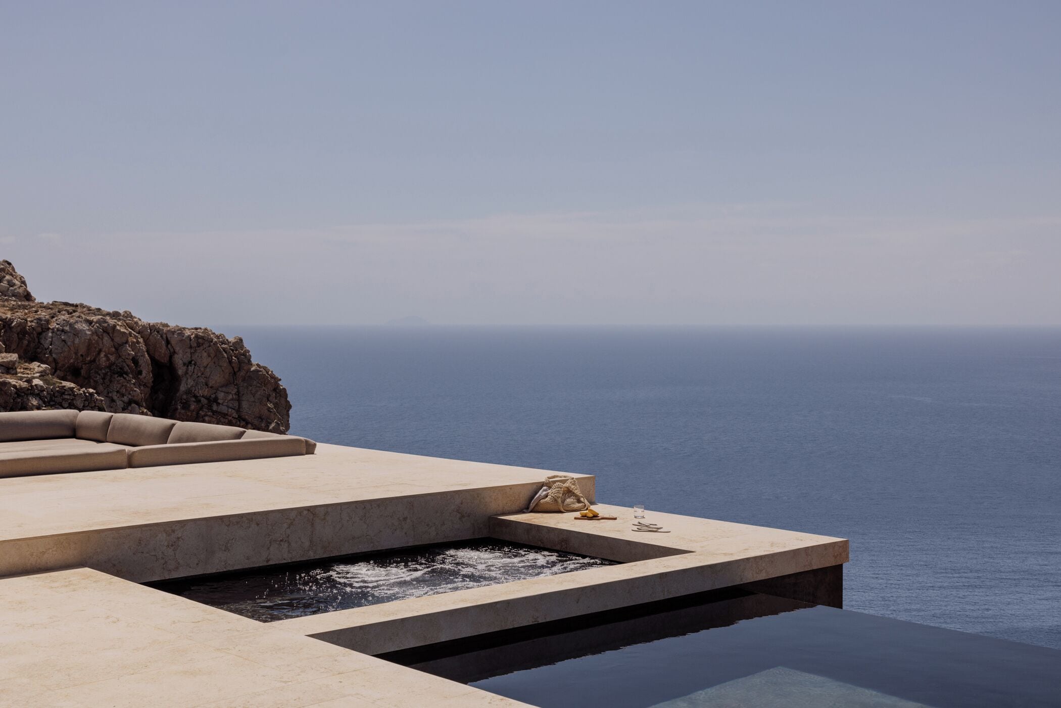 A modern infinity pool with a clear view of the ocean, a seating area with cushions, and a small table with items on it.