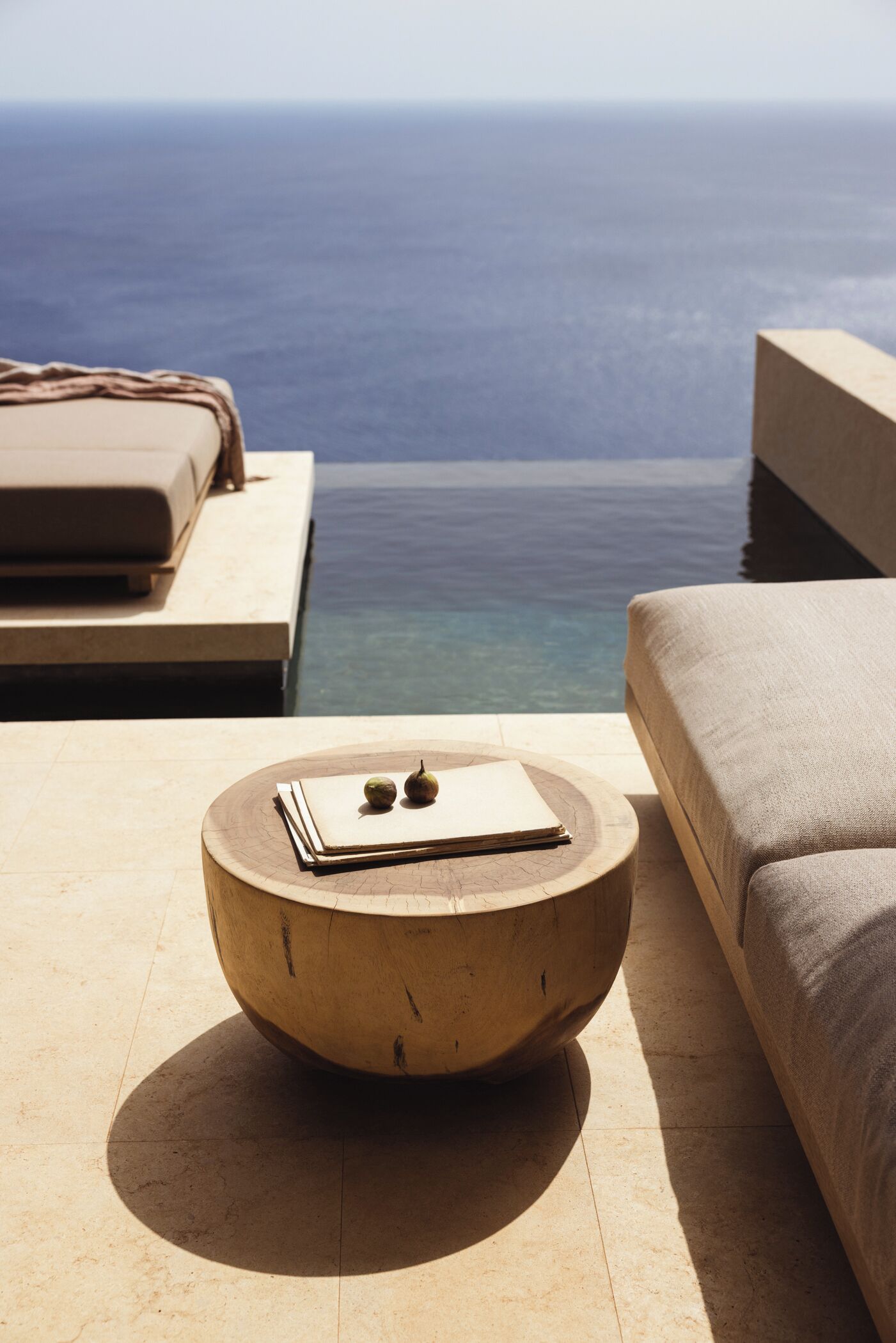 A round wooden table with a book and two apples, placed on a tiled floor near a pool with a view of the ocean.