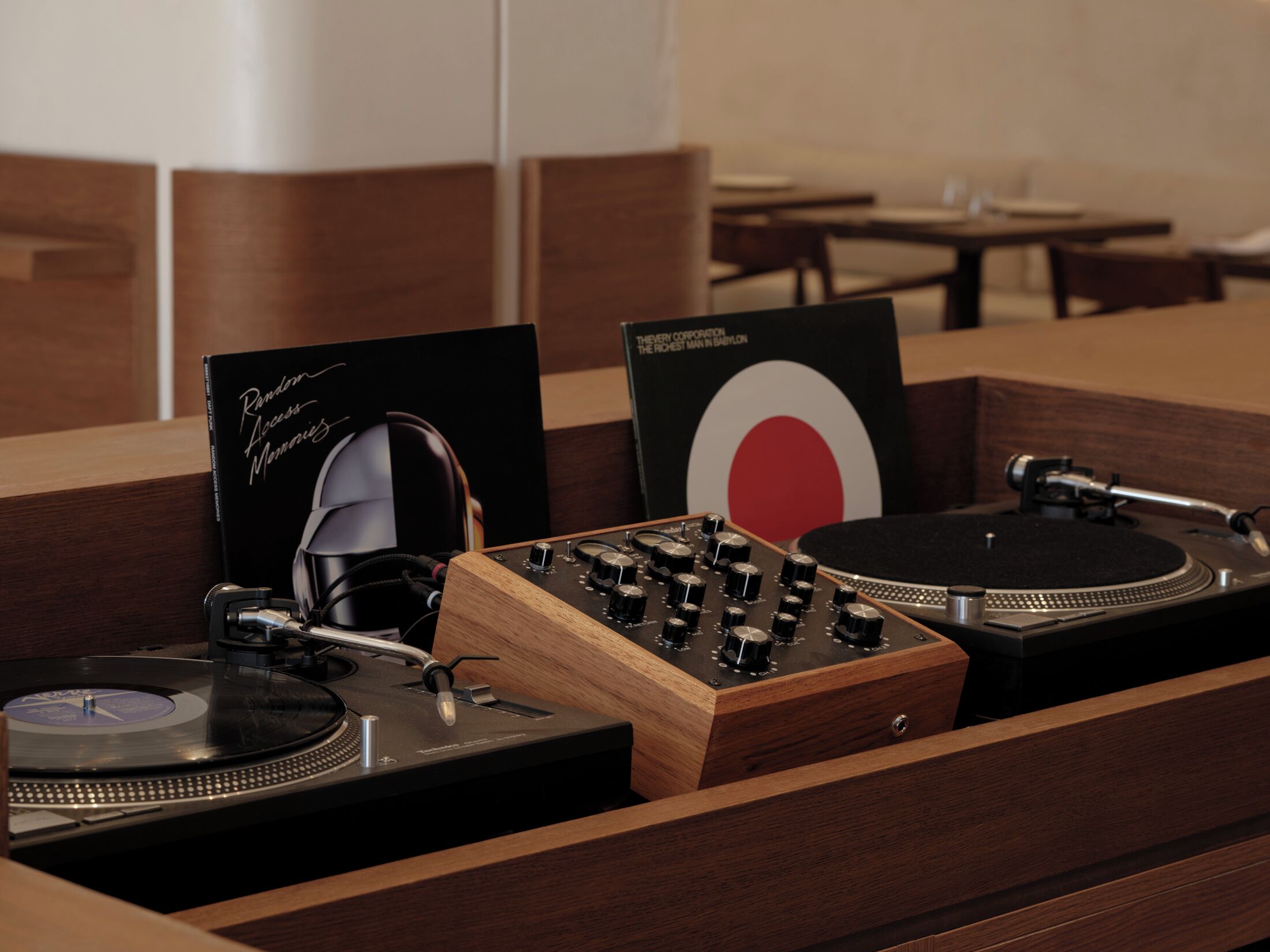 Une table de DJ en bois, équipée de platines et de matériel audio, un panneau noir avec du texte blanc, et un motif circulaire rouge et blanc.