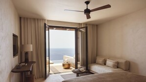 A hotel room with a bed, a desk, a lamp, a ceiling fan, and a view of the ocean. - Gundari (Folegandros)