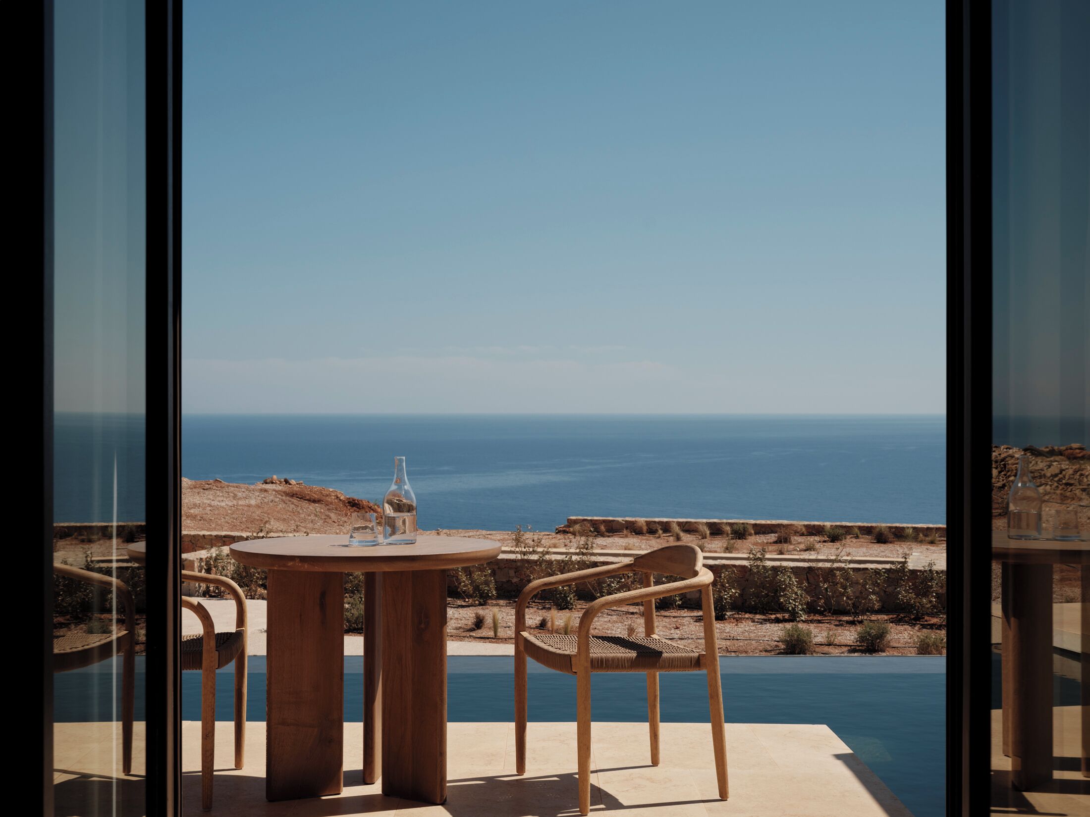 A terrace with a table and two chairs overlooking a pool and the sea.