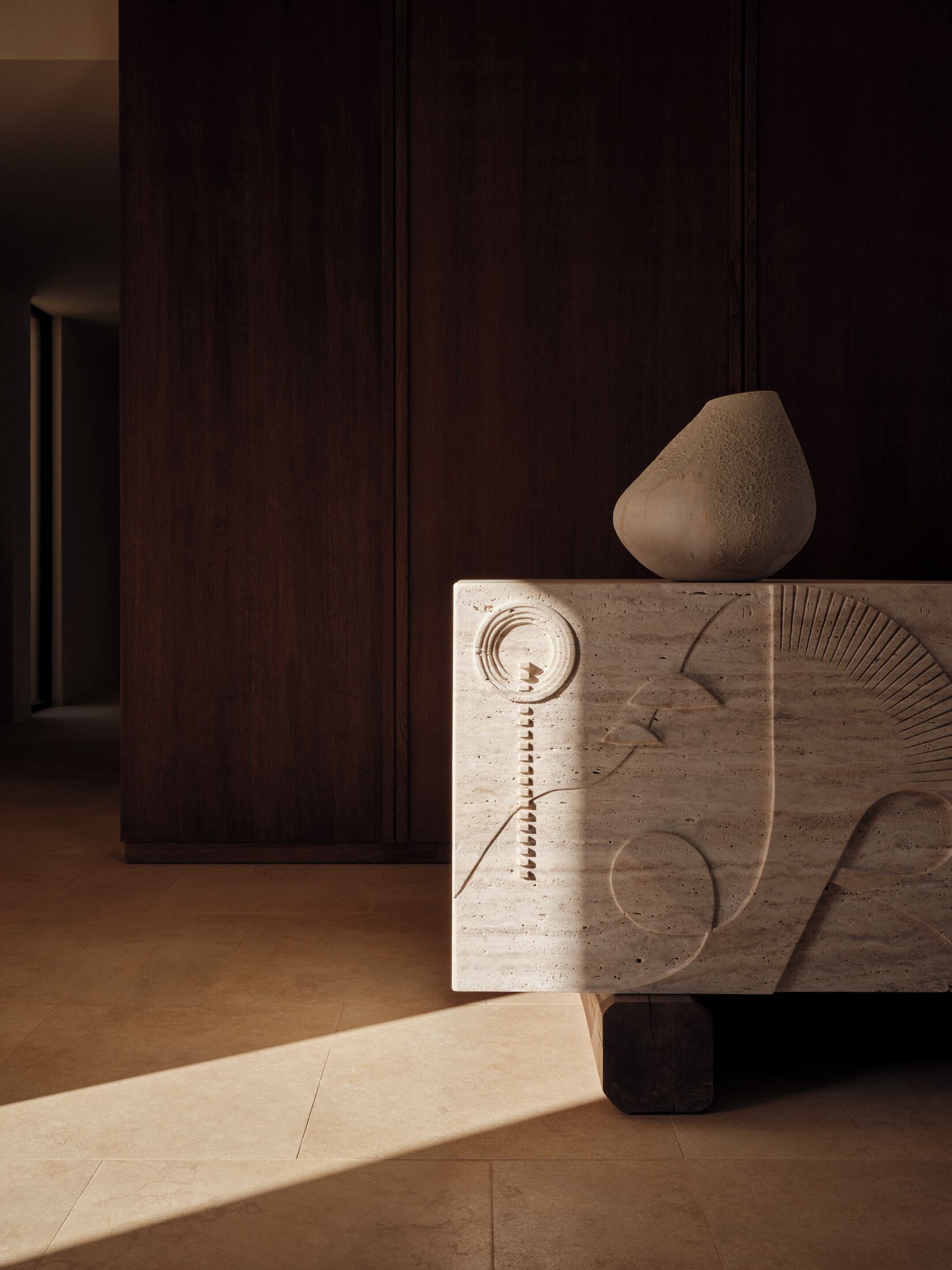 Ein schlichter Raum mit einem Holzschrank, einem skulpturalen Objekt darauf und einer Stehlampe, die einen Schatten wirft.