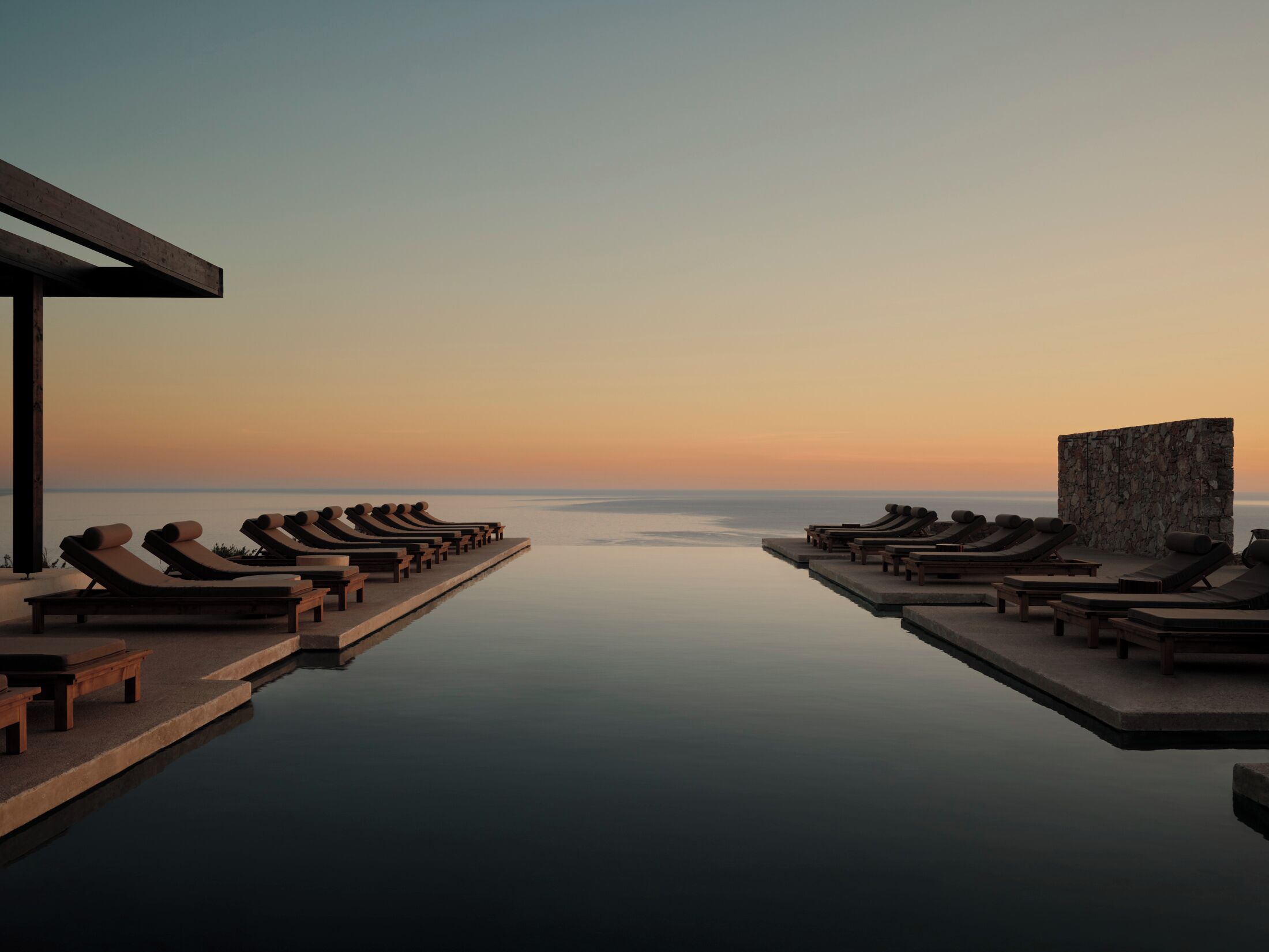 A serene infinity pool with lounge chairs and a stone wall, overlooking a calm sea at sunset.