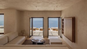 A spacious room with a large sliding glass door leading to a balcony with a view of the sea. There is a sofa, a coffee table, and a wooden cabinet. - Gundari (Folegandros)
