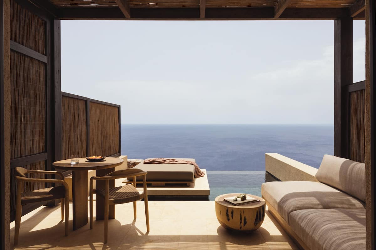 A room with a view of the sea, a round wooden table, two chairs, a sofa, and a small round table with a book and a glass.