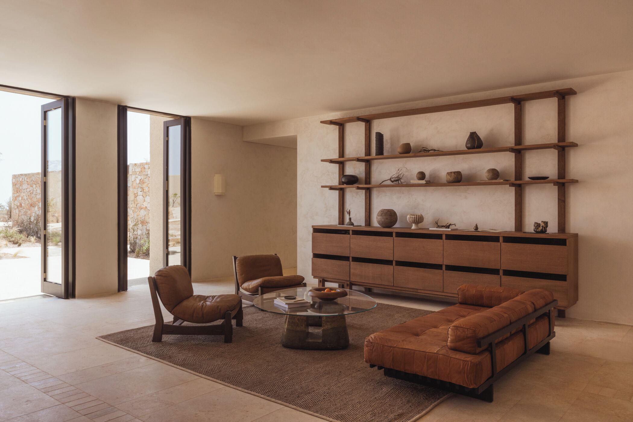 A modern living room with a glass coffee table, leather sofas, and a wooden shelving unit against the wall.