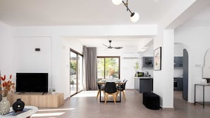A modern living room with a flat-screen TV, a dining area with a table and chairs, a kitchen area with cabinets and a countertop, and a ceiling fan.