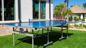 An outdoor ping pong table in front of a building with large windows and a thatched-roof structure in the background.