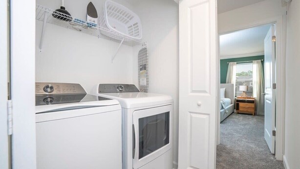 A laundry area with a washing machine and dryer, a drying rack, and a door leading to a bedroom with a bed and a nightstand.