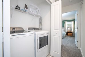Un área de lavandería con lavadora y secadora, un tendedero y una puerta que da a un dormitorio con cama y mesita de noche.