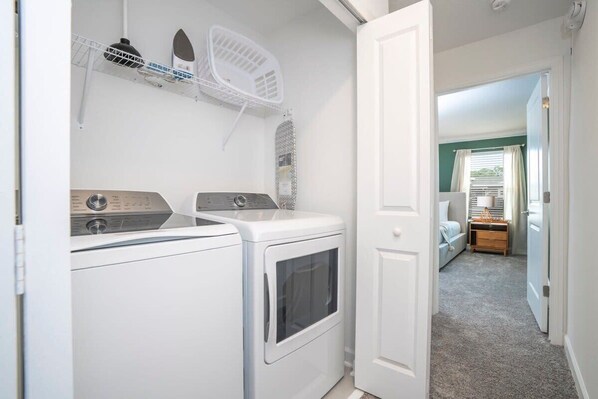 A laundry area with a washing machine and dryer, a drying rack, and a door leading to a bedroom with a bed and a nightstand.