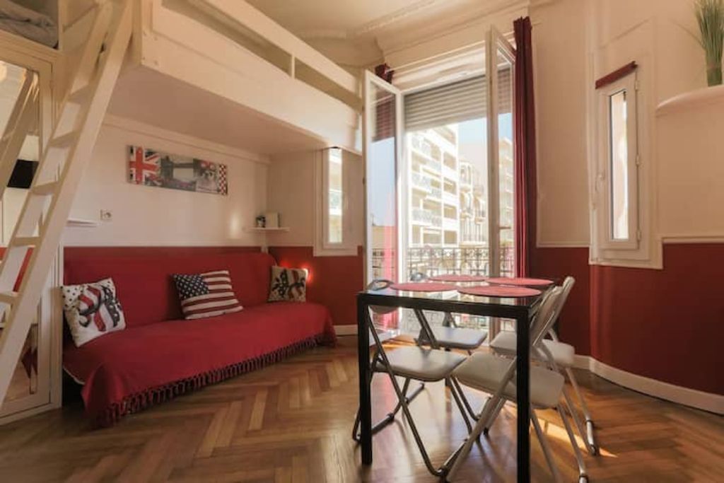 Una habitació acollidora amb un sofà vermell, una taula de menjador amb caires i una finestra amb cortines.