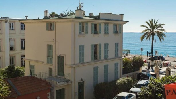 Un bâtiment situé sur la côte, doté de nombreuses fenêtres et de balcons, et jouxtant une plage bordée de palmiers, sous un ciel bleu et dégagé.