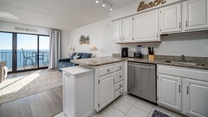 A modern kitchen with white cabinets, a granite countertop, and a view of the sea through a sliding glass door.