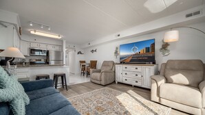 A modern living room with a blue sofa, beige armchairs, a patterned rug, and a kitchen in the background.