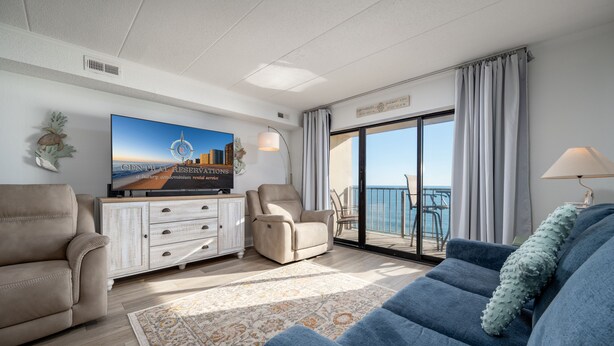 A modern living room with a flat-screen TV, a sofa, a chair, and a view of the ocean through a sliding glass door.