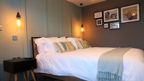 A neatly made bed with white linens and striped pillows, a bedside table with a lamp, and framed artwork on the wall. - Diglis Basin Apartment (Worcester)