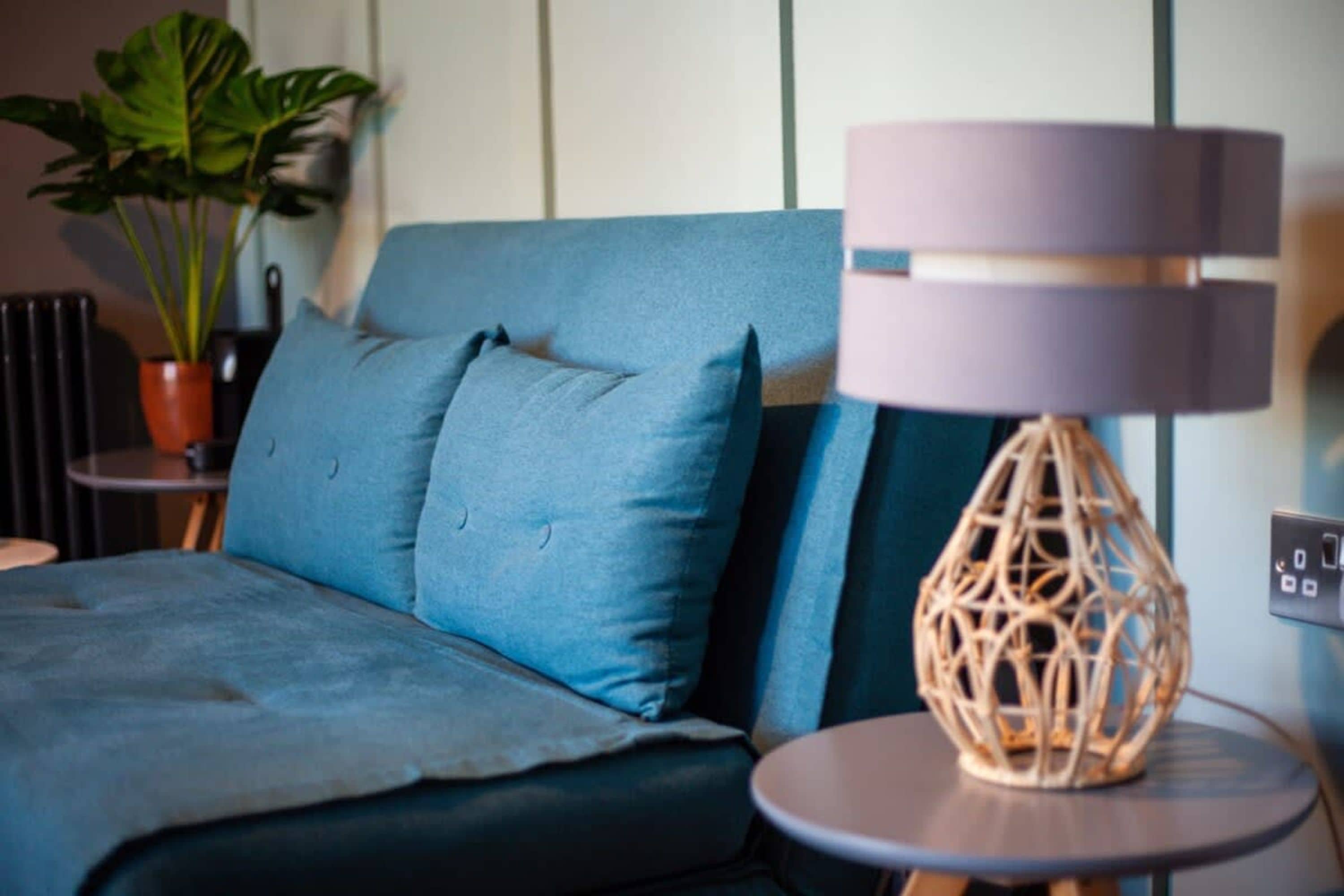 A modern bedroom with a blue sofa, a round table with a woven lamp, and a potted plant.