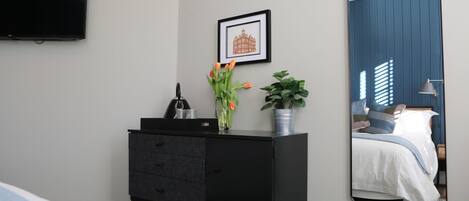 A bedroom with a black dresser, a framed picture on the wall, a potted plant, and a mirror reflecting a bed.