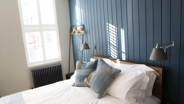 A bedroom with a bed, pillows, a nightstand, a lamp, and a window with blinds. - Hop Market - Ensuite Bedroom (Worcester)