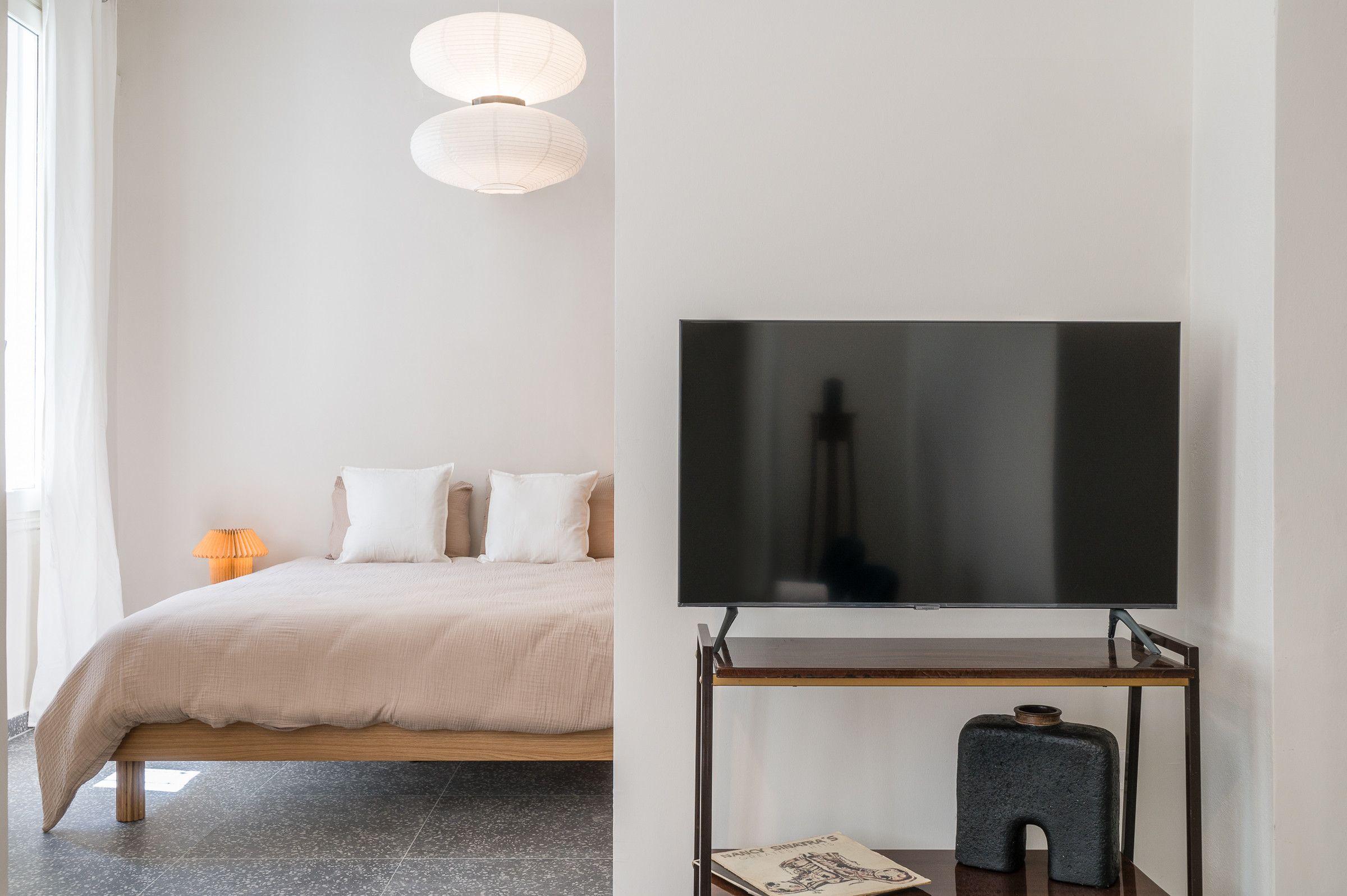 Un dormitorio con cama, almohadas, una lámpara de noche y un televisor sobre un soporte.