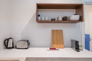 A kitchen with a wooden shelf holding plates, glasses, and a mug. There is a toaster, a kettle, and a cutting board with cherries on the counter.