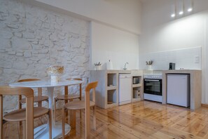A compact kitchen with a stone wall, white tiles, wooden chairs, a dining table, a dishwasher, an oven, and a refrigerator.