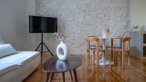 Un salón con pared de piedra, un televisor de pantalla plana, un sofá blanco, una mesa redonda de madera con sillas y un jarrón blanco con flores.