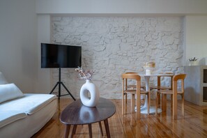 A living room with a stone wall, a flat-screen TV, a white sofa, a round wooden table with chairs, and a white vase with flowers.