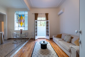 A modern living room with a glass dining table, a sofa, and a large window with curtains. - Charlie – Timeless Charm & Modern Comfort (Athens)