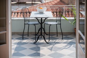 Un balcon avec une table et deux chaises, un pot de fleurs et une vue sur un immeuble aux tuiles en terre cuite.