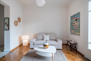 Un salon moderne avec un canapé gris clair, une table basse blanche, un luminaire suspendu et un tableau abstrait aux couleurs vives accroché au mur.