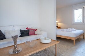 A minimalist bedroom with a wooden bed, a white bedside table, and a white sofa with cushions.