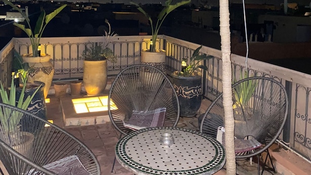 Terraço no telhado com área de estar ao ar livre, incluindo cadeiras de metal e uma mesa redonda, cercado por plantas em vasos e um grande guarda-sol.