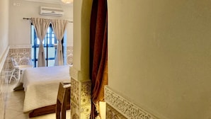 Quarto de hotel com cama, uma mesa pequena, uma cadeira, janela com cortinas e um painel decorativo na parede.