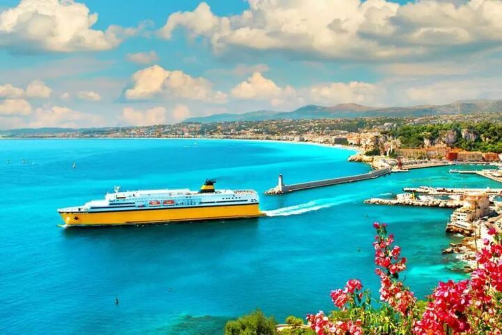 A cruise ship in a harbor with clear blue waters, a lighthouse, and a coastal town in the background.
