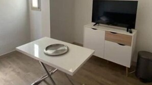 A small, sparsely furnished room with a white table, a television on a white cabinet, and a window providing natural light. - Le Port - Cosy studio perfectly located (Nice)