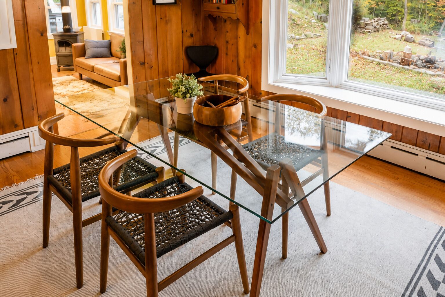 A dining area with a glass table and wooden chairs, a potted plant, and a view of the outdoors.