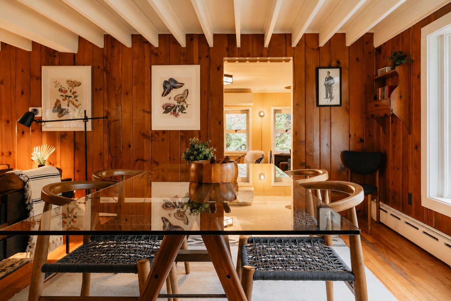 Een kamer met houten lambrisering, een glazen eettafel, gevlochten stoelen en ingelijste kunstwerken aan de wanden.